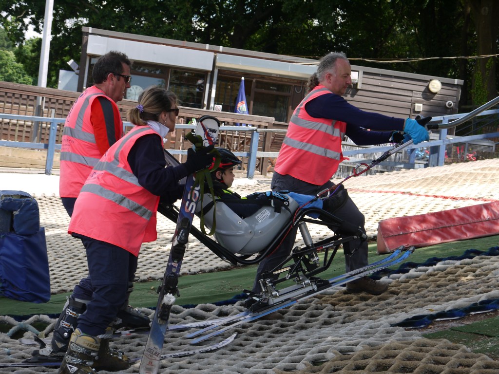 Boy in sit ski being helped onto the button lift by 3 Guides.